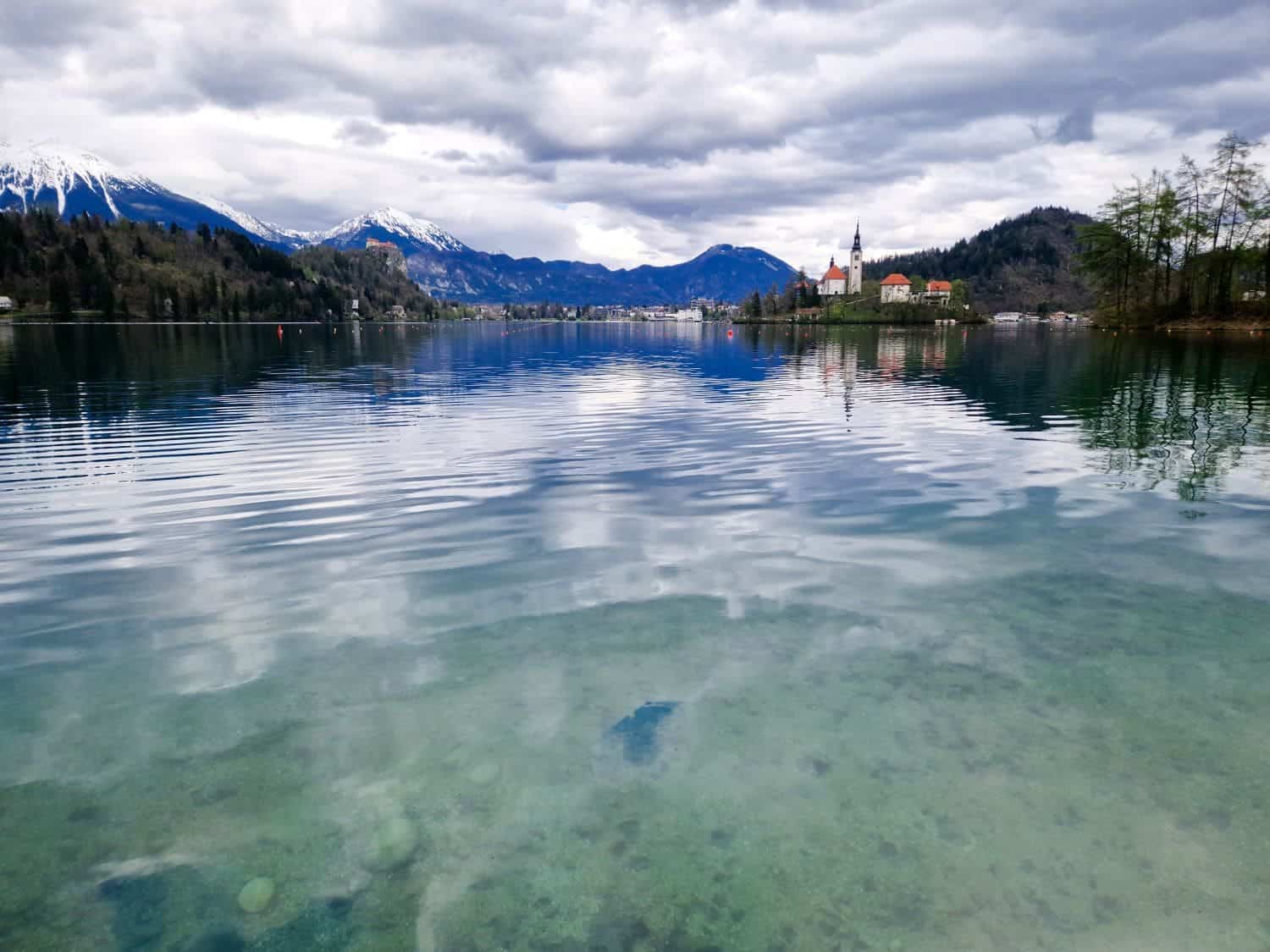 Dolomites In April: Perfect 2-Day Itinerary + What To Expect 52 A serene lake with clear water reflecting a cloudy sky and distant mountains. An island with a church and buildings is in the middle, surrounded by trees and hills. The water's foreground is transparent, showing the lakebed.