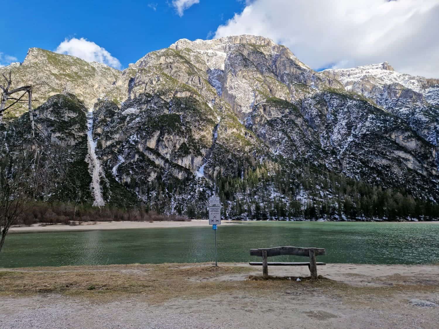 Dolomites In The Summer: Tips And Tricks For Visiting In 2025 15 A serene mountain landscape featuring a calm lake in the foreground and snow-capped peaks under a blue sky with clouds. A wooden bench faces the lake, and a signpost stands nearby, adding a human element to the natural scene.