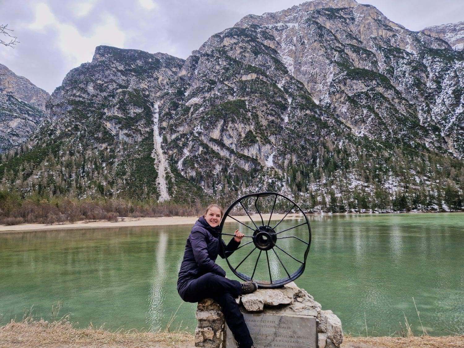 Dolomites In April: Perfect 2-Day Itinerary + What To Expect 25 A person in a black jacket sits on a rock near a scenic lake, holding a large wheel sculpture. Snow-dusted mountains and evergreen trees frame the background, under a cloudy sky.