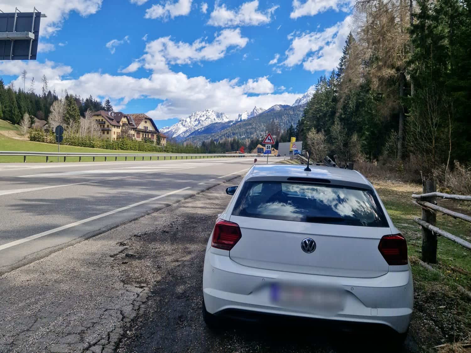 Dolomites In April: Perfect 2-Day Itinerary + What To Expect 57 A white car is parked on the side of a scenic road under a blue sky with scattered clouds. Snow-capped mountains and evergreen trees line the background. A few buildings are visible on the left.