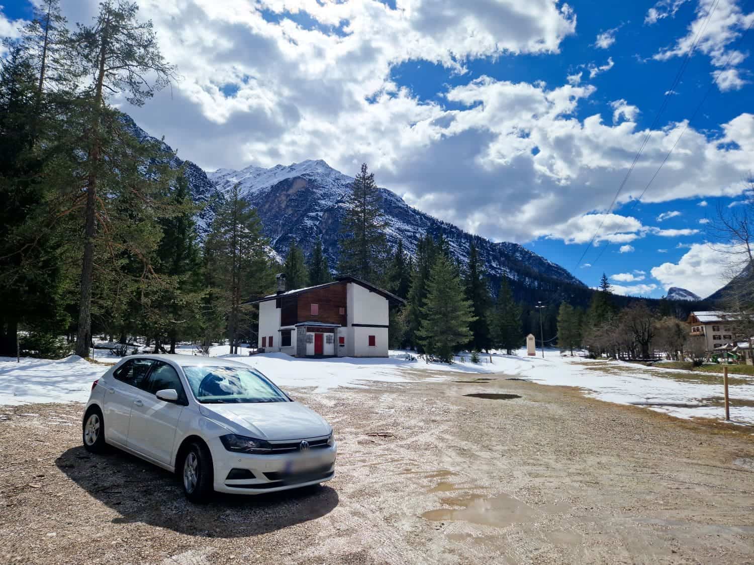 Dolomites In April: Perfect 2-Day Itinerary + What To Expect 18 A white car is parked on a dirt path near a house surrounded by snow. Tall pine trees and a mountain range are in the background under a partly cloudy sky.