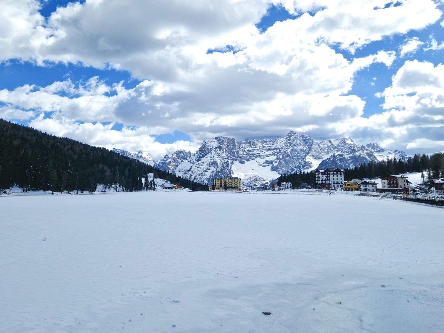 Dolomites In April: Perfect 2-Day Itinerary + What To Expect 19 A snowy landscape featuring a frozen lake in the foreground, with buildings nestled among trees on the right. towering snow-covered mountains in the background under a partly cloudy blue sky.