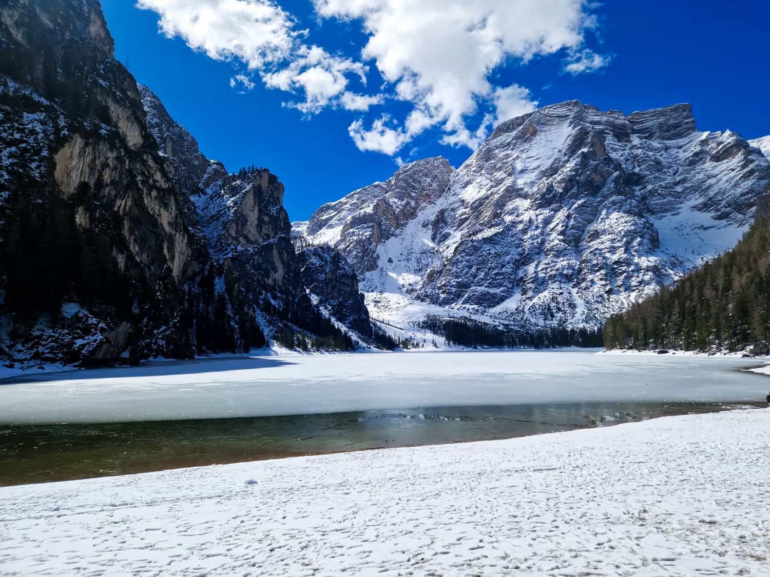 Dolomites In April: Perfect 2-Day Itinerary + What To Expect 6 Snow-covered mountains surround a partially frozen lake under a vibrant blue sky with scattered clouds. A thin layer of snow covers the ground in the foreground. Pine trees line the right side of the landscape.