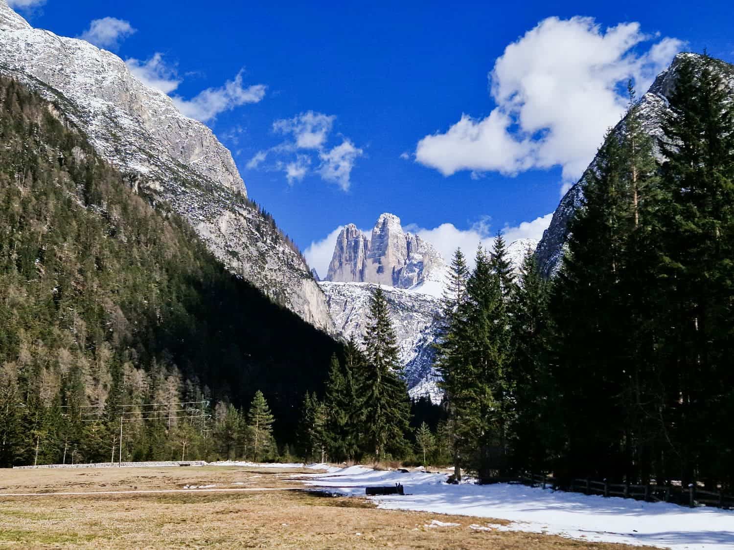 Easy Dolomites Hiking Itinerary: 5 days in Cortina d’Ampezzo 7 A scenic view of a mountainous landscape with snow-capped peaks under a blue sky. Pine trees frame the foreground, with patches of snow on the ground. Fluffy clouds float above, adding contrast to the vibrant scene.