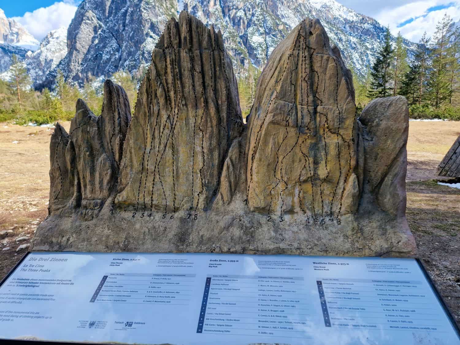 Dolomites In April: Perfect 2-Day Itinerary + What To Expect 22 A rock display resembling a mountain range with numbered climbing routes is set against a backdrop of real mountains. A sign in front provides information about the climbing paths. Snow-capped peaks and trees are visible in the background.