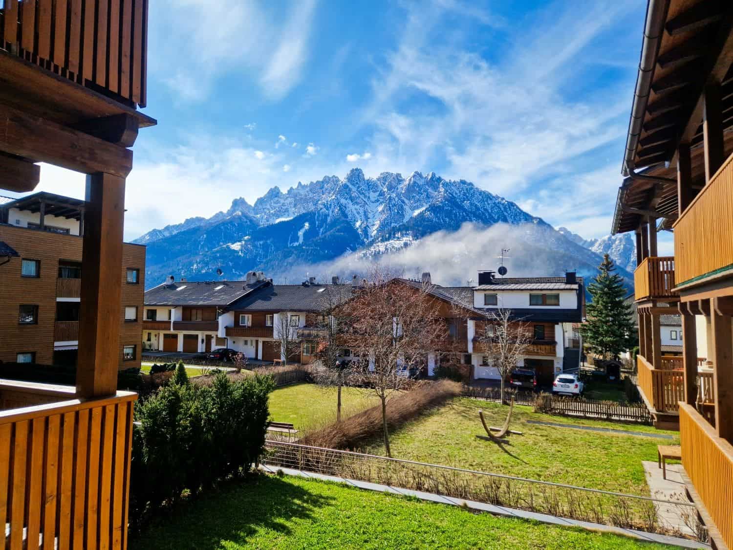 Dolomites In April: Perfect 2-Day Itinerary + What To Expect 58 Mountain view from a wooden balcony with residential houses in front. Snow-capped peaks under a partly cloudy sky create a scenic backdrop. A grassy yard with a swing is visible between the buildings.