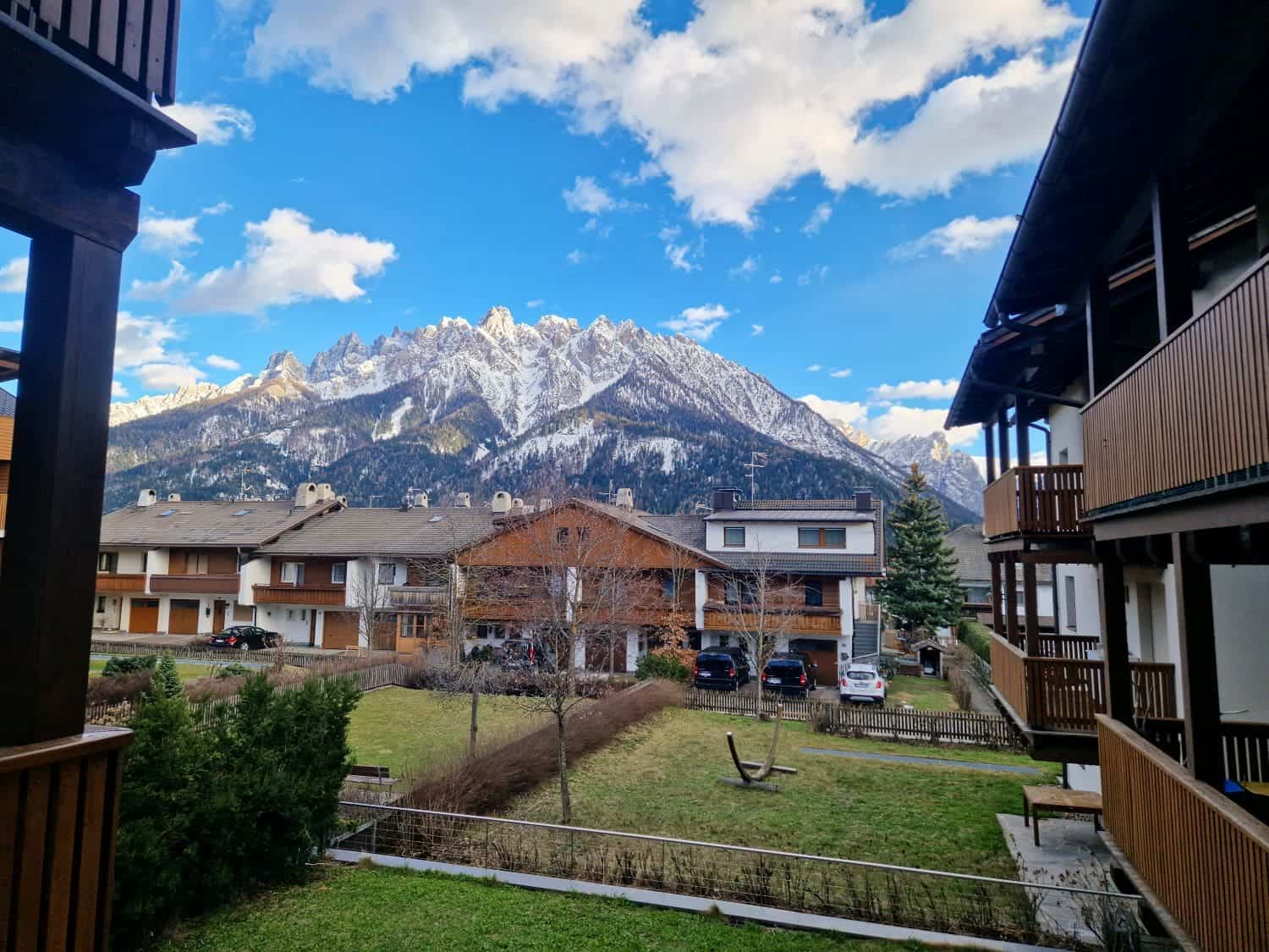 16 Best Places To Visit In The Dolomites In Summer 60 View of a snowy mountain range under a partly cloudy sky, framed by wooden residential buildings. A grassy yard with bare trees is visible, along with a few parked cars in front of the houses.