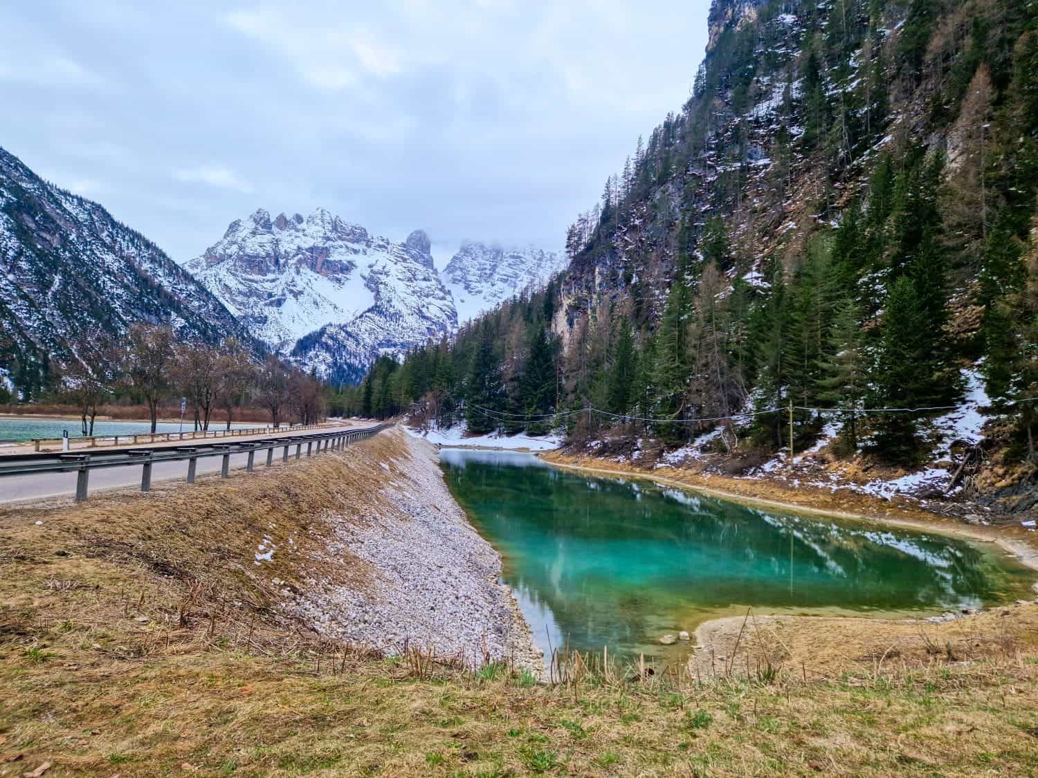 Easy Dolomites Hiking Itinerary: 5 days in Cortina d’Ampezzo 6 A serene turquoise pond sits beside a mountain road with a wooden guardrail. Snow-dusted trees line the surrounding rugged terrain, and snow-covered peaks rise in the background under a cloudy sky.