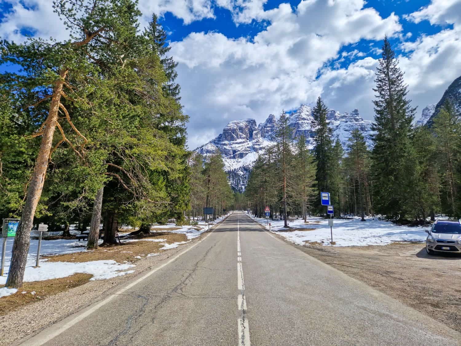 Dolomites In April: Perfect 2-Day Itinerary + What To Expect 60 A straight road in a snowy forest leads towards distant snow-covered mountains under a partly cloudy blue sky. Trees line the roadside, and a car is parked in a small lot to the right. Signs and patches of melting snow are visible.