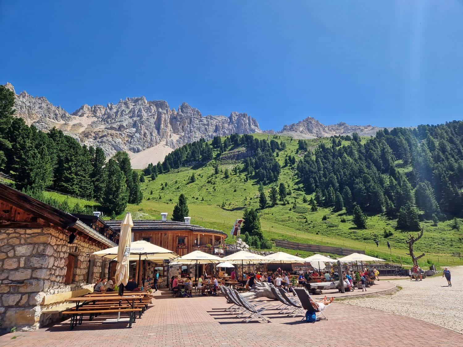 16 Best Places To Visit In The Dolomites In Summer 61 Outdoor seating area with umbrellas and lounge chairs on a sunny day, surrounded by green hills and pine trees. A rocky mountain forms the backdrop under a clear blue sky. People are relaxing and enjoying the scenery.