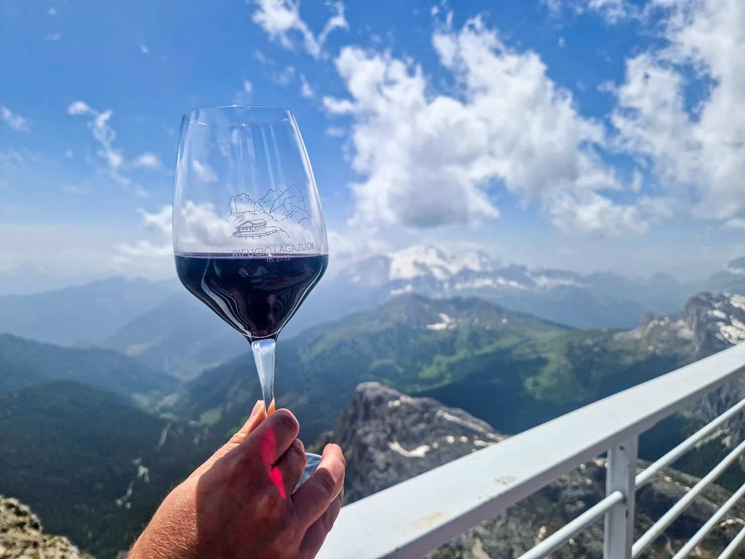 Easy Dolomites Hiking Itinerary: 5 days in Cortina d’Ampezzo 30 A person holds a glass of red wine against a backdrop of a mountainous landscape with lush green valleys and snow-capped peaks under a blue sky with scattered clouds. The glass features a mountain logo.