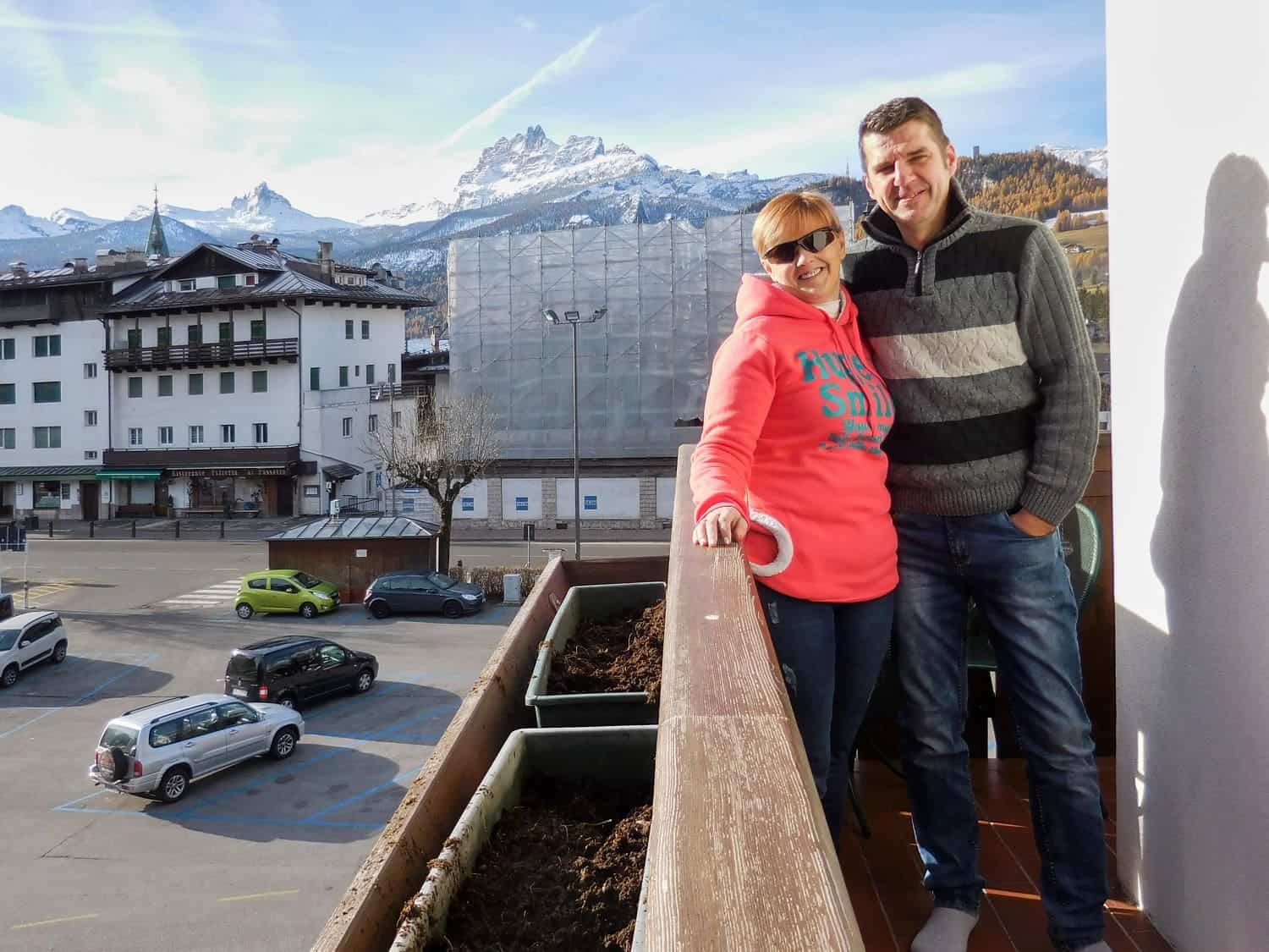 16 Best Places To Visit In The Dolomites In Summer 59 A man and woman stand on a balcony smiling. The woman wears a pink hoodie and sunglasses, and the man wears a gray sweater. Behind them, there are mountains, buildings, and parked cars under a clear blue sky.