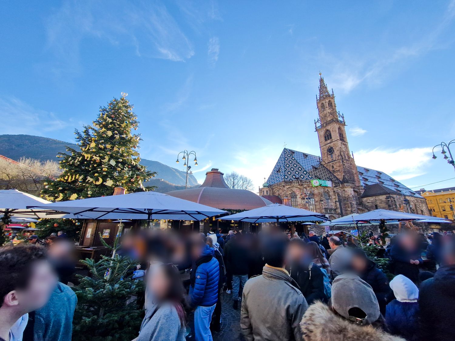 How to Get to Cortina d’Ampezzo From Key Destinations 7 A crowded Christmas market fills a town square with people standing beneath white vendor umbrellas and evergreen decorations. A large decorated Christmas tree stands to the left, while a historic church with a patterned roof and tall bell tower rises behind the stalls under a clear blue sky. The image shows a festive winter atmosphere in a European city center.