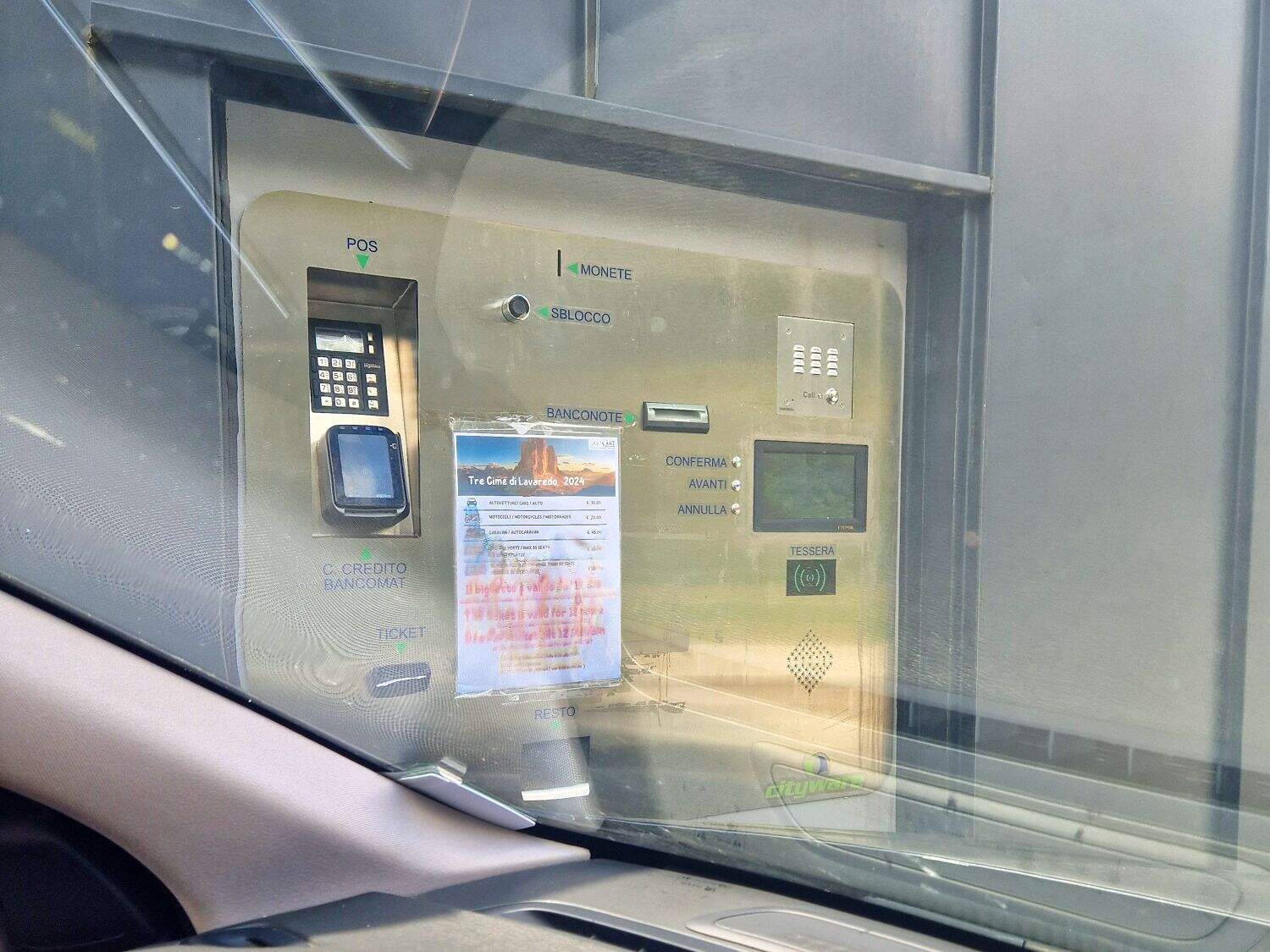 All About the Rifugio Auronzo Toll Road: Gateway to Tre Cime 7 A parking payment machine viewed from a car window. It includes card and cash slots, a keypad, and a screen. A partially obscured poster is taped to it. The surroundings appear to be an indoor parking facility.
