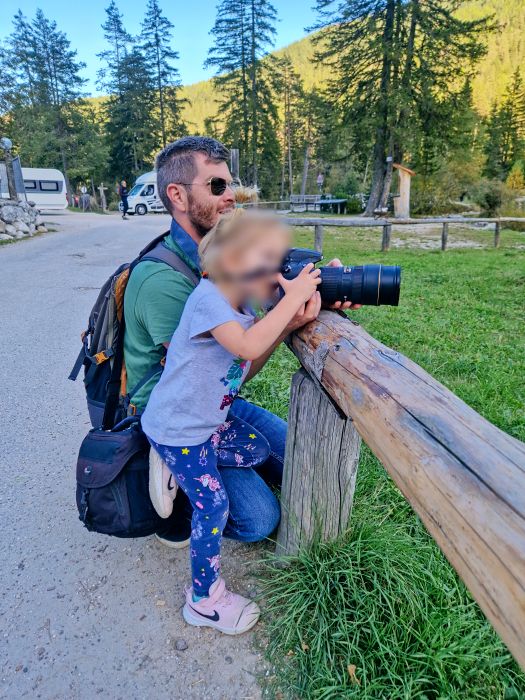 15 Best Photo Spots in the Dolomites: MAP + Complete Guide 23 An adult kneels next to a child, helping them hold a large camera on a wooden fence in a park. Trees and a parked camper van are visible in the background under a sunny sky.