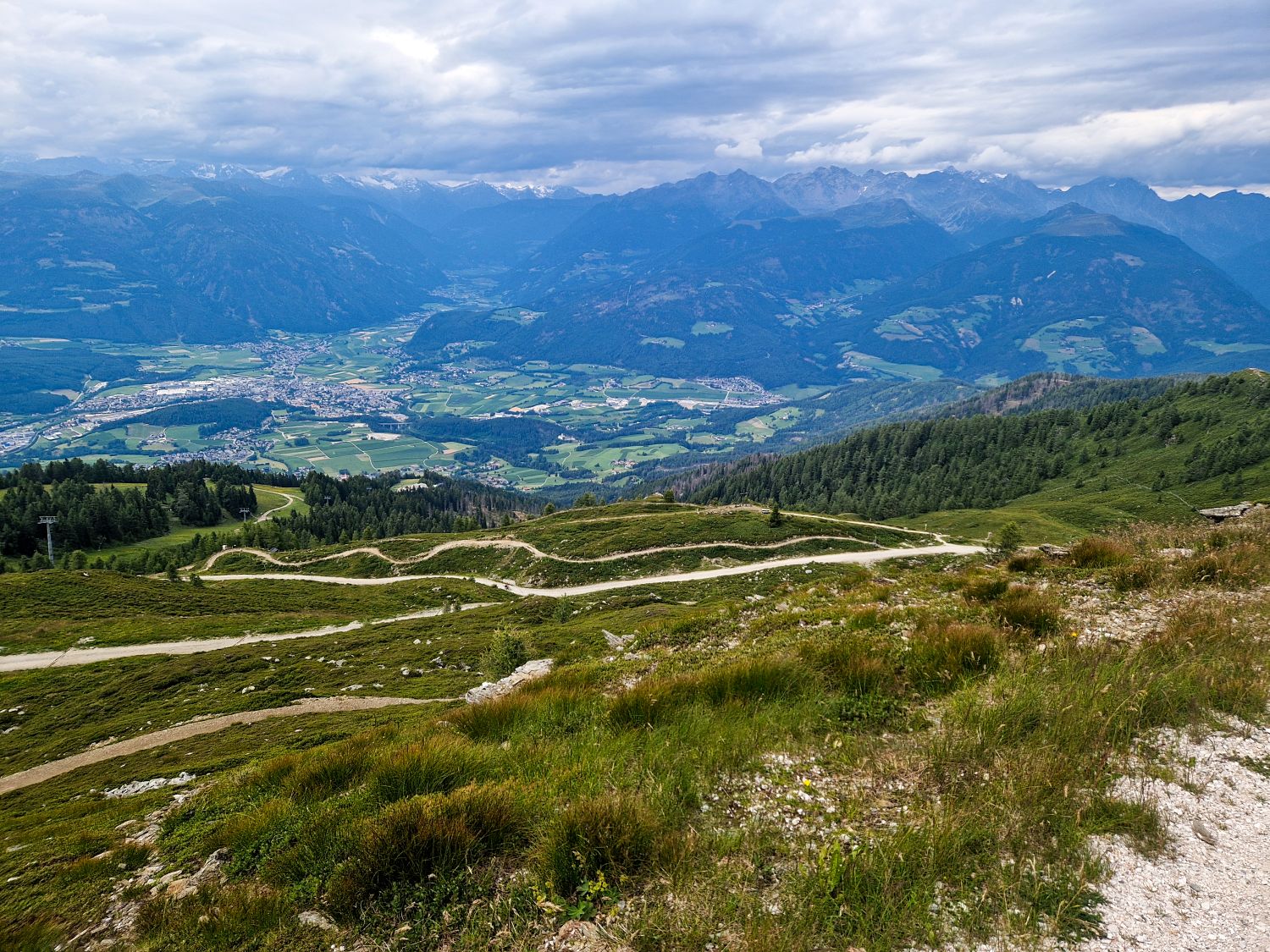 11 Hotels With The Best Views Of The Dolomites: Worth Every Penny 4 A winding mountain road snakes through grassy hills, overlooking a lush valley with towns and fields, surrounded by distant, forested mountains under a cloudy sky.