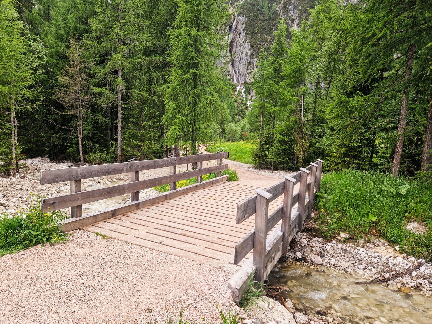 Hiking To The Cascate Del Pisciadù In The Dolomites: Plus a Surprise Waterfall! 12 A wooden footbridge crosses a small stream in a forested area, surrounded by lush green trees and rocky terrain. A dirt path leads onto the bridge, with mountains visible in the background.