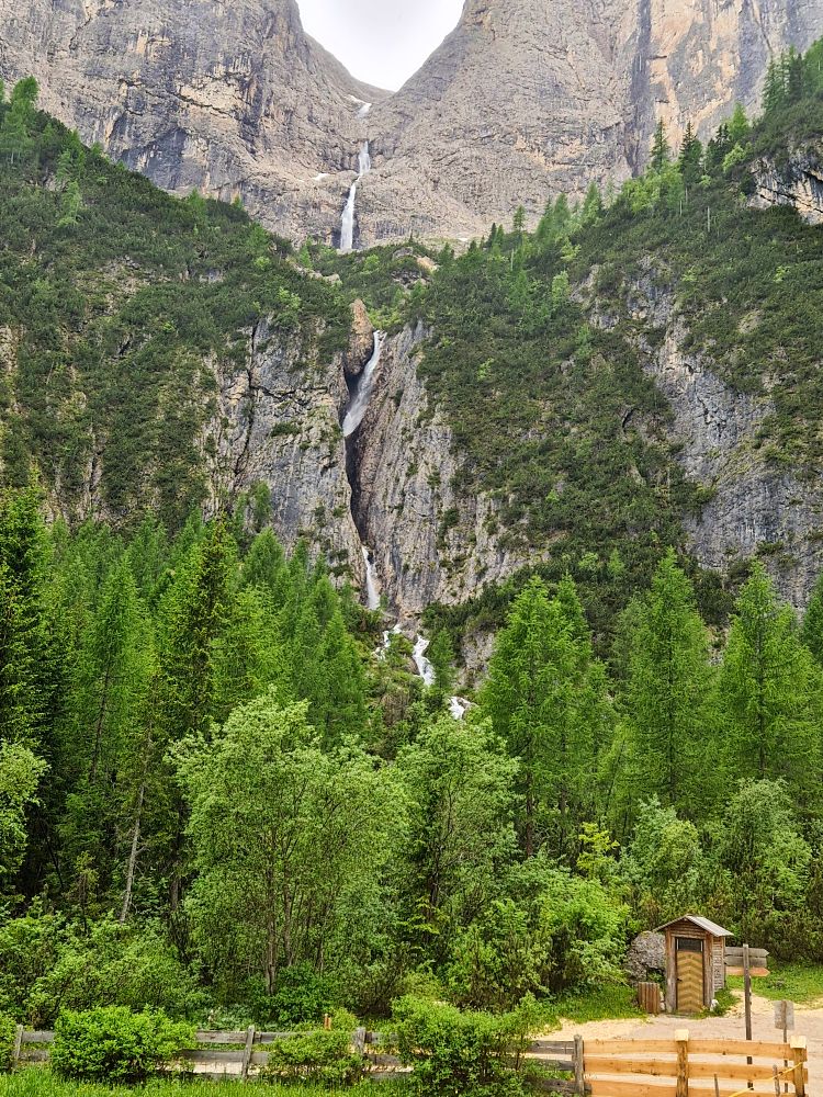 Hiking To The Cascate Del Pisciadù In The Dolomites: Plus a Surprise Waterfall! 2 A tall, narrow waterfall cascades down a steep, rocky mountain surrounded by dense green pine trees and a wooden shed in the foreground.
