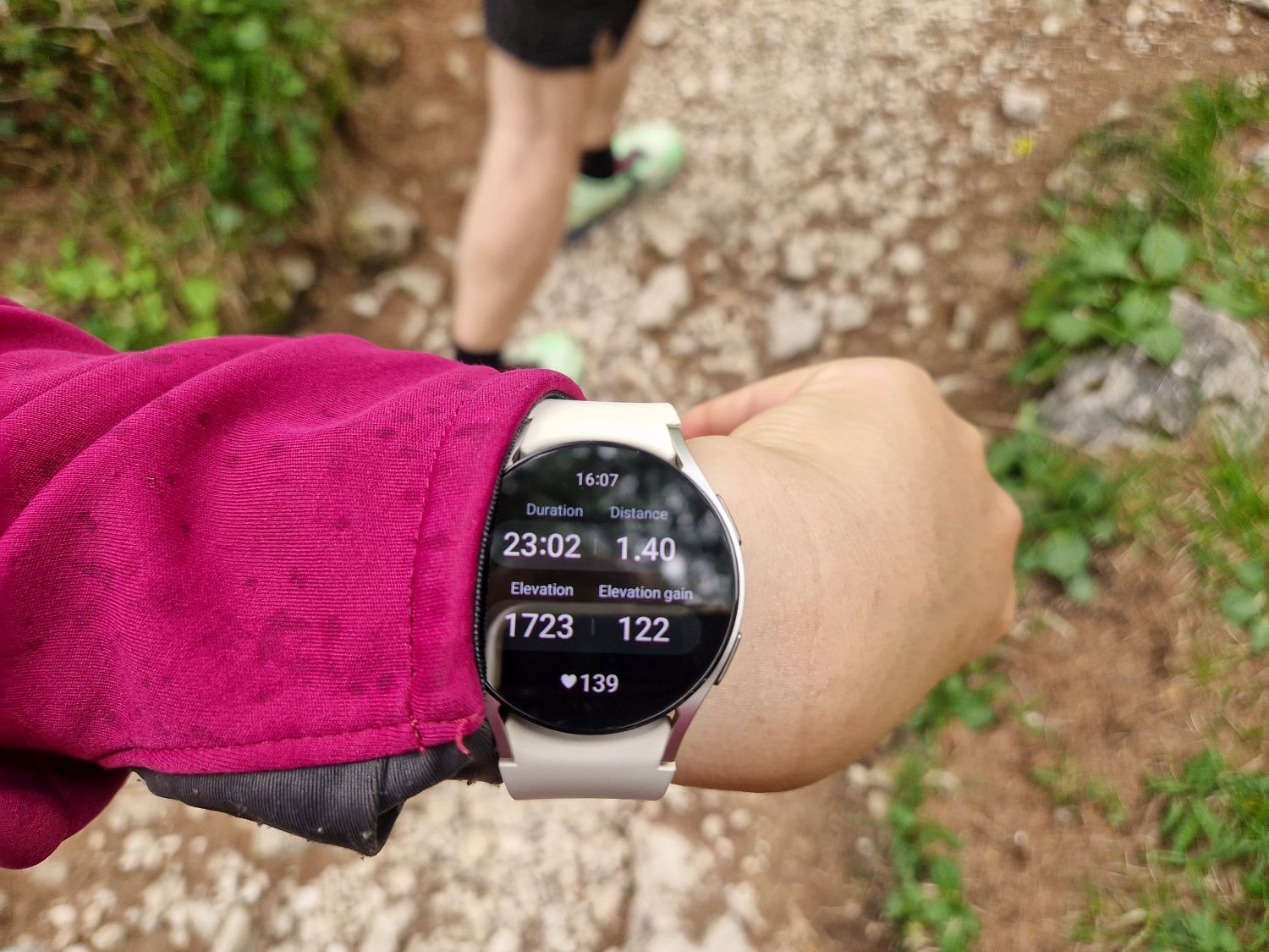 Hiking To The Cascate Del Pisciadù In The Dolomites: Plus a Surprise Waterfall! 3 A person wearing a maroon jacket checks their smartwatch, displaying hiking stats: 23:02 duration, 1.40 km distance, 1723 m elevation, 122 m elevation gain, and 139 bpm heart rate. Another hiker walks ahead on a rocky trail.