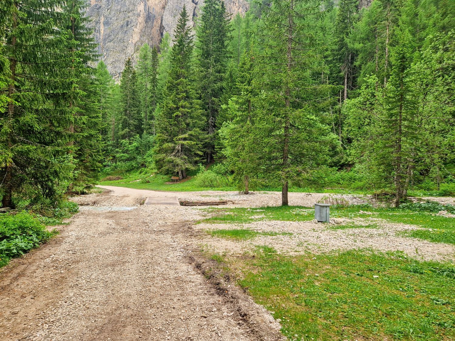 Hiking To The Cascate Del Pisciadù In The Dolomites: Plus a Surprise Waterfall! 25 A dirt path winds through a lush green forest with tall pine trees, a small gravel clearing, and a metal trash can on the right side under cloudy daylight.