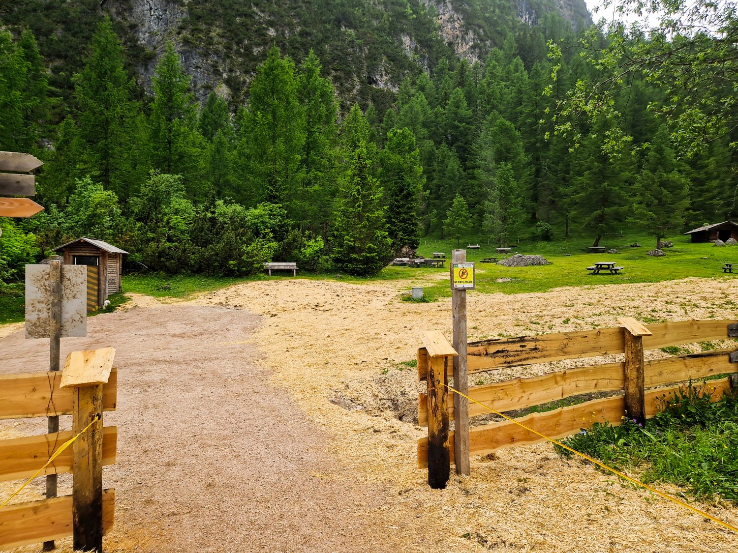 Hiking To The Cascate Del Pisciadù In The Dolomites: Plus a Surprise Waterfall! 13 A fenced area with wood shavings on the ground opens to a grassy clearing surrounded by dense green trees, with several benches and picnic tables scattered throughout the scene.