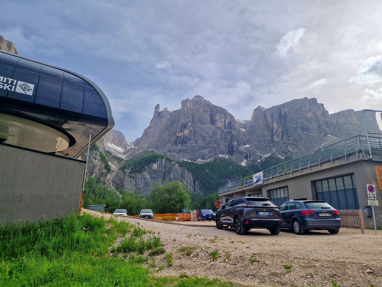 Hiking To The Cascate Del Pisciadù In The Dolomites: Plus a Surprise Waterfall! 38 Cars parked near a modern ski lift station, with dramatic rocky mountains and green forests in the background under a partly cloudy sky.