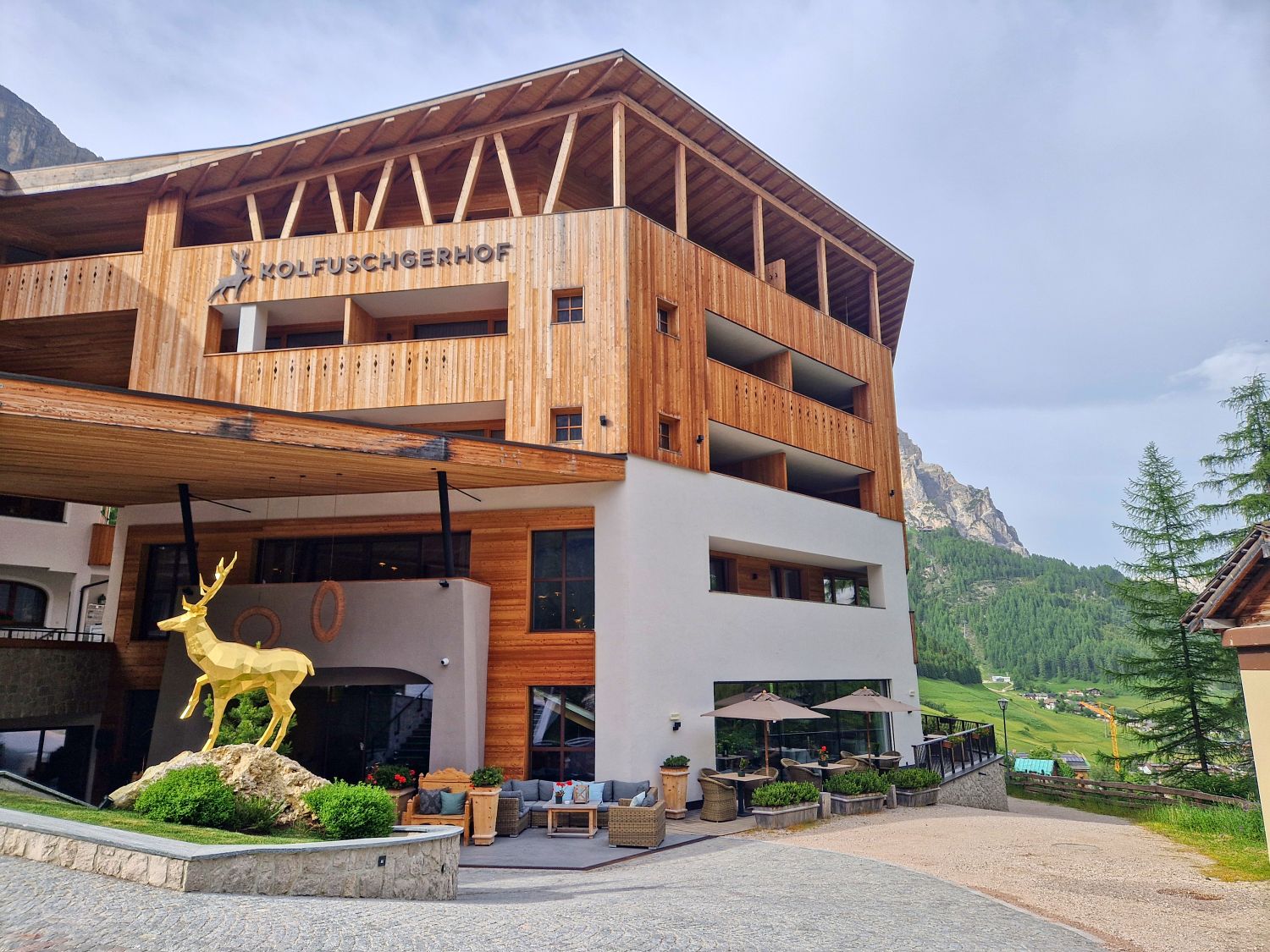 Hiking To The Cascate Del Pisciadù In The Dolomites: Plus a Surprise Waterfall! 47 A modern wooden hotel with the name "Kolfuschgerhof" on the facade. There's a large golden deer statue on a rock near the entrance, with outdoor seating and mountains visible in the background.