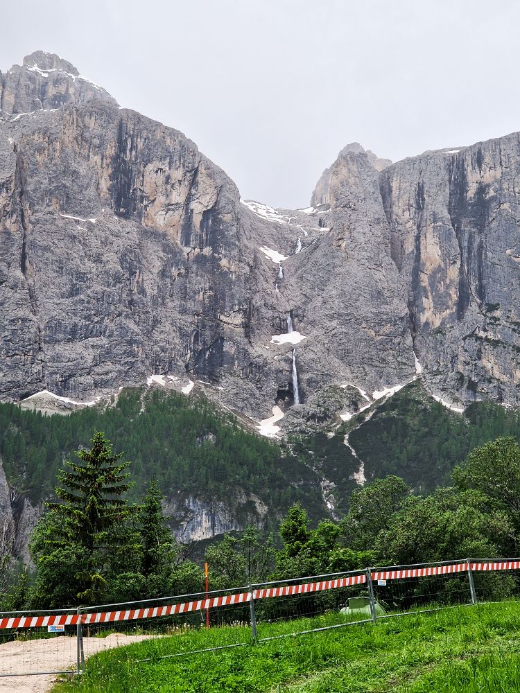 Hiking To The Cascate Del Pisciadù In The Dolomites: Plus a Surprise Waterfall! 7 A tall, rocky mountain with patches of snow and a narrow waterfall cascading down the center, bordered by a green forest and a striped barrier in the foreground.