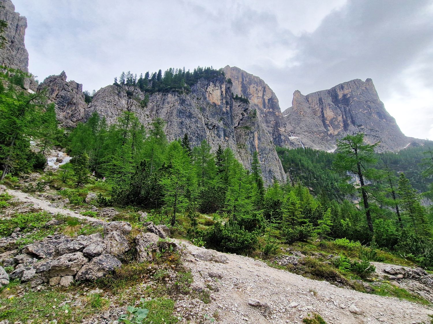 Hiking To The Cascate Del Pisciadù In The Dolomites: Plus a Surprise Waterfall! 35 A rocky mountain trail winds through green pine trees with steep, rugged cliffs rising in the background under a cloudy sky.