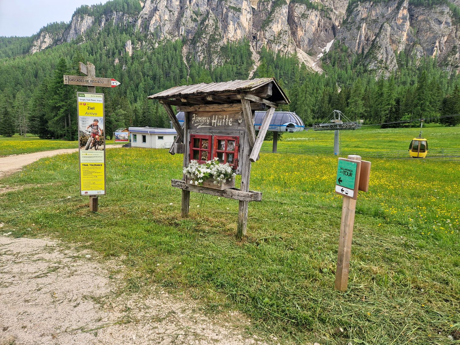 Hiking To The Cascate Del Pisciadù In The Dolomites: Plus a Surprise Waterfall! 40 A rustic wooden sign with flowers stands in a grassy field near a yellow info board, a green post, and a gondola; forested mountains and a ski lift are visible in the background.