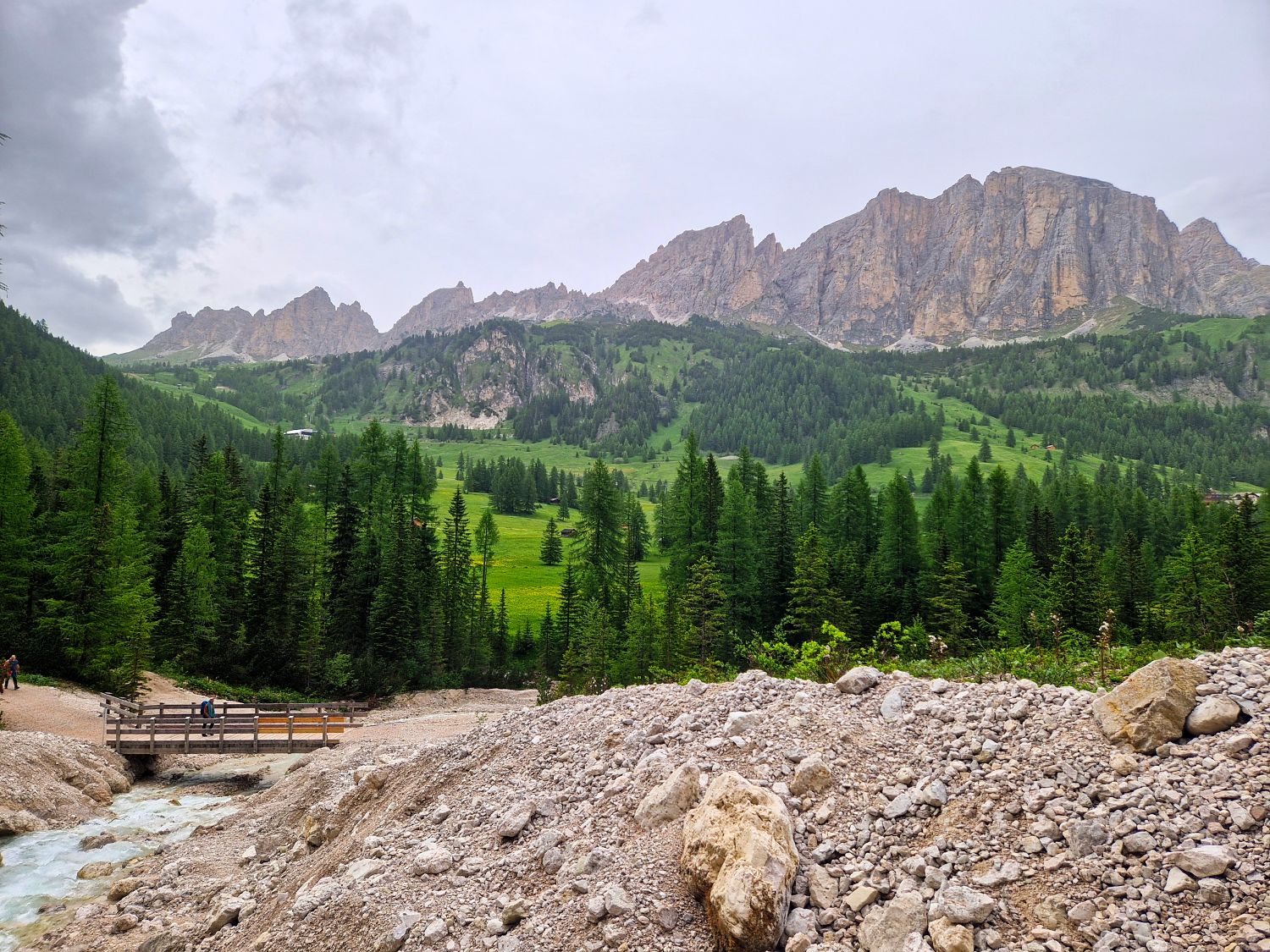 Hiking To The Cascate Del Pisciadù In The Dolomites: Plus a Surprise Waterfall! 30 A rocky foreground leads to a small wooden bridge over a stream, surrounded by dense pine trees and green hills, with tall, rugged mountains in the background under a cloudy sky.