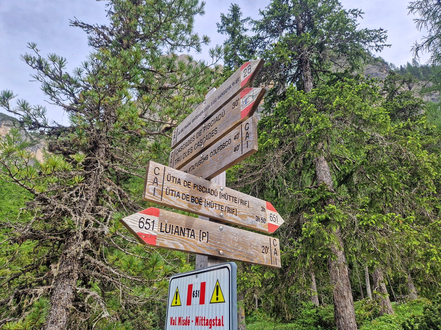 Hiking To The Cascate Del Pisciadù In The Dolomites: Plus a Surprise Waterfall! 31 Wooden trail signs with red and white markings point in multiple directions among tall green trees, indicating hiking routes and distances in a forested mountain area.