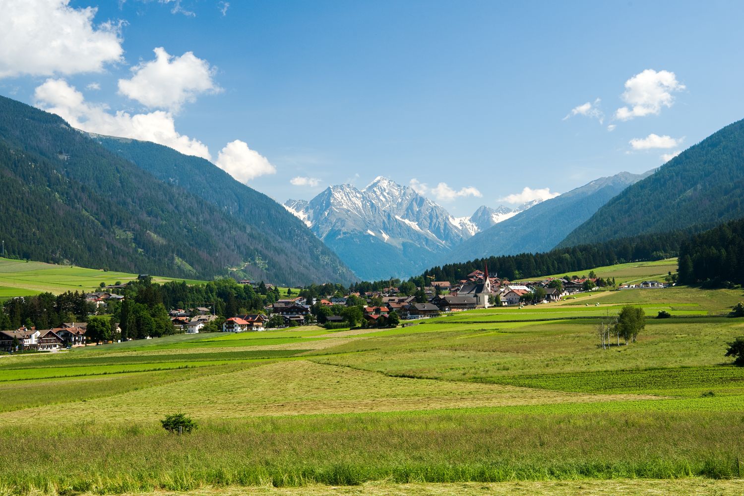11 Hotels With The Best Views Of The Dolomites: Worth Every Penny 7 A picturesque village lies in a wide green valley surrounded by forested hills and snow-capped mountains under a bright blue sky with clouds.