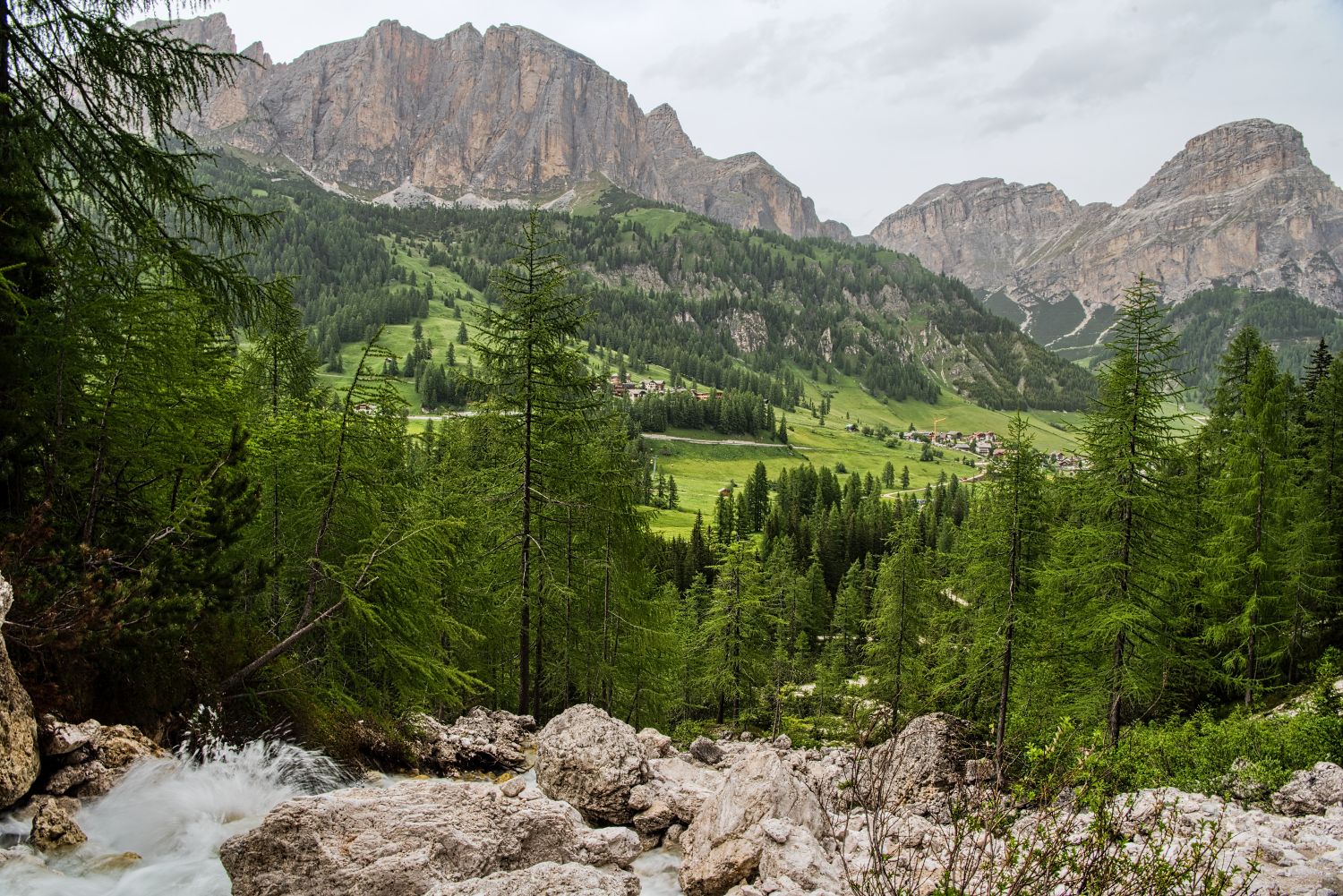 Hiking To The Cascate Del Pisciadù In The Dolomites: Plus a Surprise Waterfall! 48 A lush, green valley with pine trees, rocky outcrops, and a small stream in the foreground, nestled beneath rugged, towering mountains under an overcast sky. Small houses are scattered in the valley.