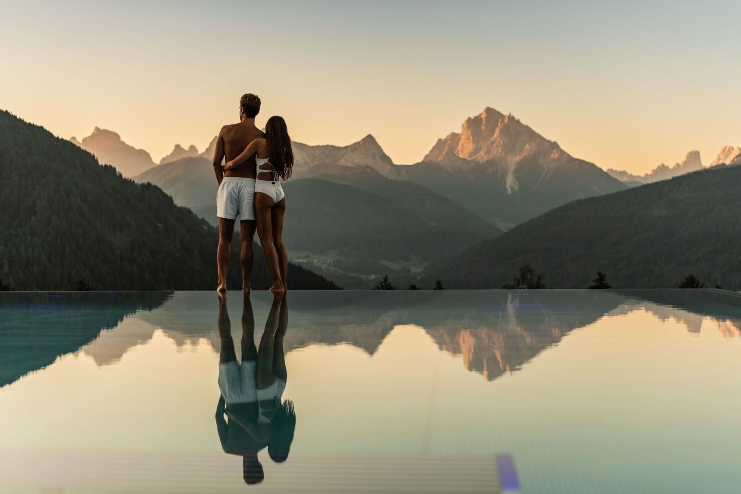 11 Hotels With The Best Views Of The Dolomites: Worth Every Penny 9 A couple in swimwear stands at the edge of an infinity pool, embracing as they look at distant mountains during sunset. The mountains and couple are reflected in the calm pool water.