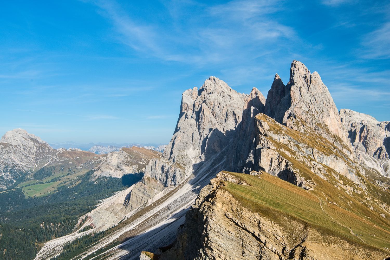 15 Best Photo Spots in the Dolomites: MAP + Complete Guide 7 Mountain landscape with jagged rocky peaks, grassy slopes, and patches of snow under a clear blue sky. Rolling hills and distant mountains are visible in the background.