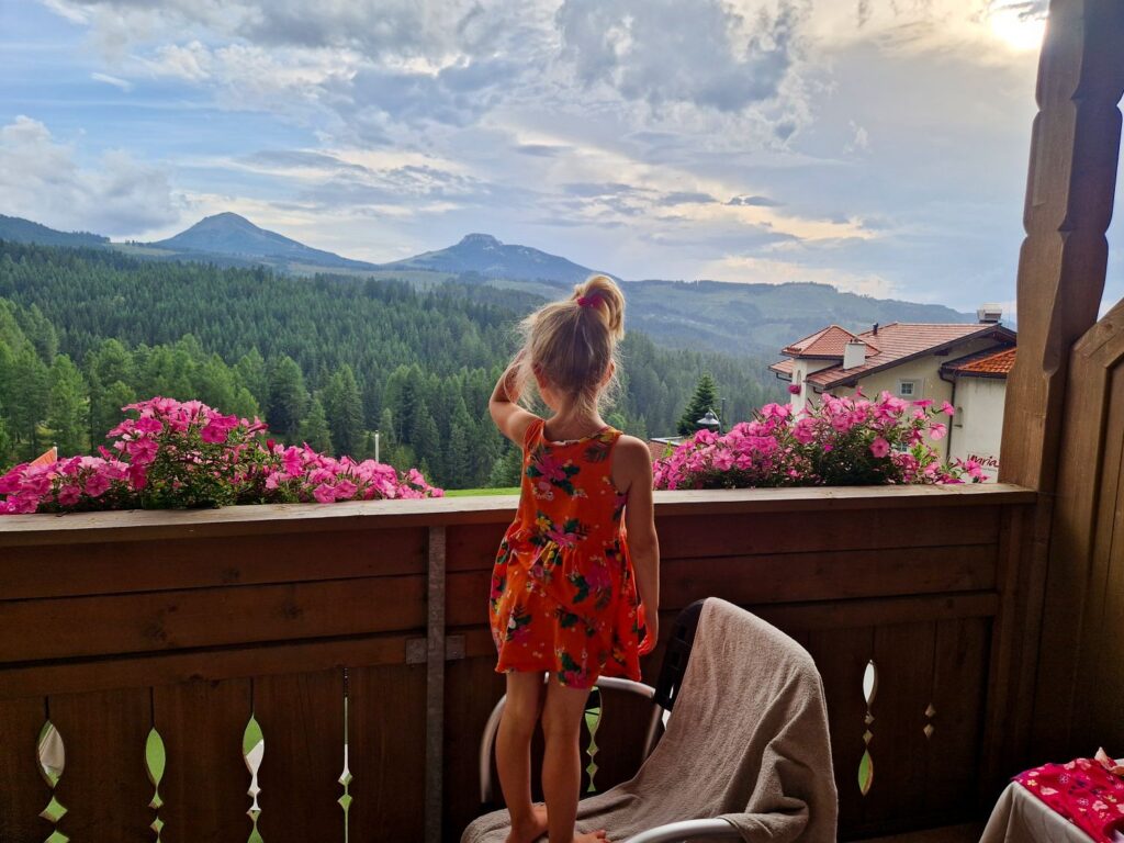 A young girl in an orange dress stands on a chair on a balcony, looking out at a scenic view of mountains, pine forests, and a cloudy sky. Pink flowers line the balcony railing.