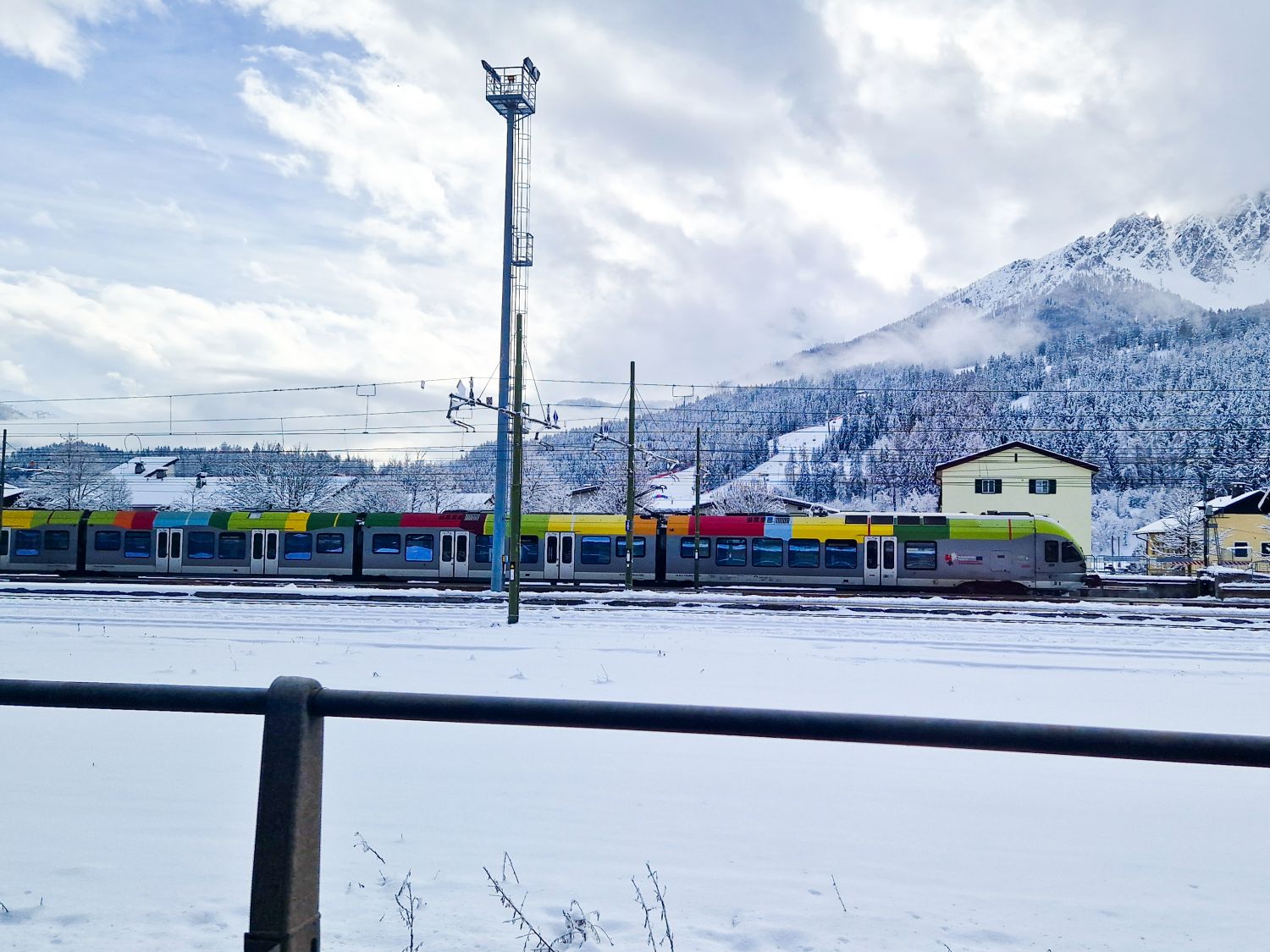 A colorful train travels through a snowy landscape, with snow-covered mountains and pine trees in the background. A yellow building stands near the tracks under a cloudy sky.