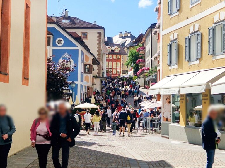 A lively, sunlit pedestrian street in a European town with colorful buildings, flower boxes, outdoor seating, and many people walking and socializing on a cobblestone path.