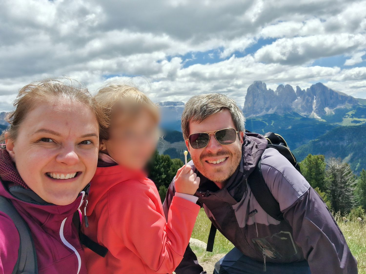 A smiling woman, a man in sunglasses, and a young child in outdoor jackets pose for a selfie on a grassy hillside with mountains and a partly cloudy sky in the background. The child's face is blurred.