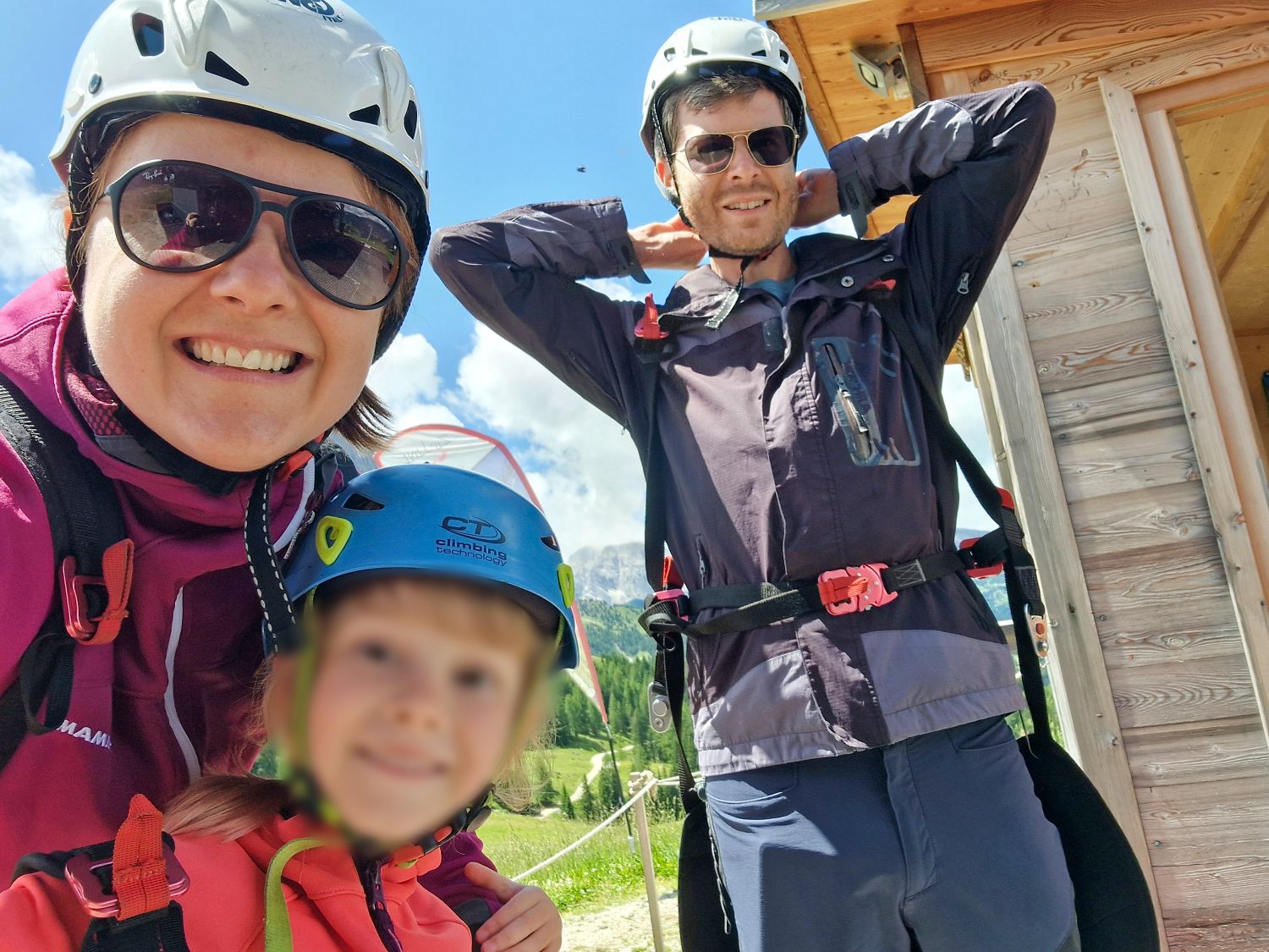A smiling family of three wearing helmets and outdoor jackets stand together outside on a sunny day, ready for an adventure. A wooden building and green landscape are visible in the background.