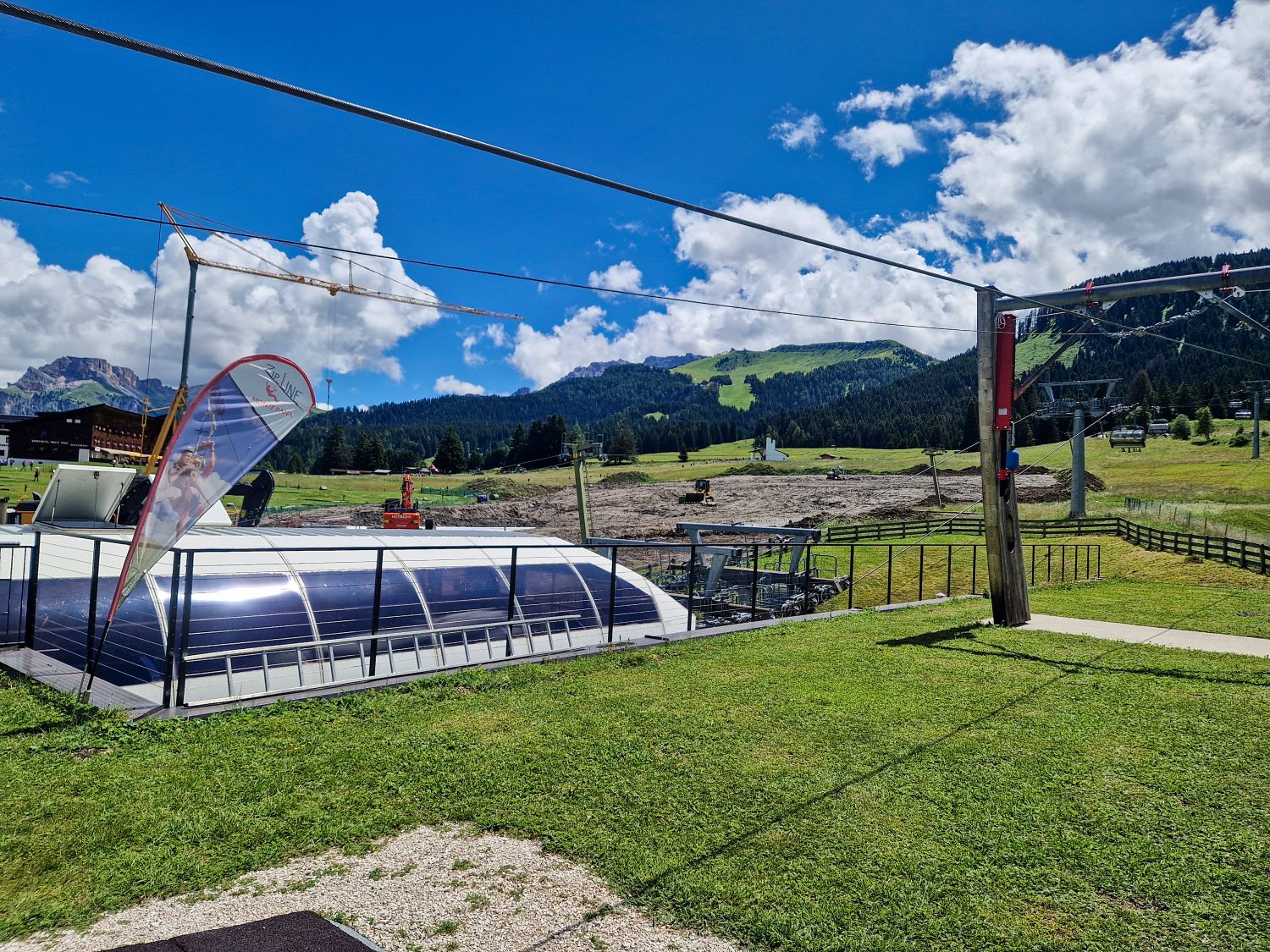 A grassy landscape with a covered chairlift station, wooden poles, and cables in the foreground. Mountains, trees, and a partly cloudy blue sky are in the background.