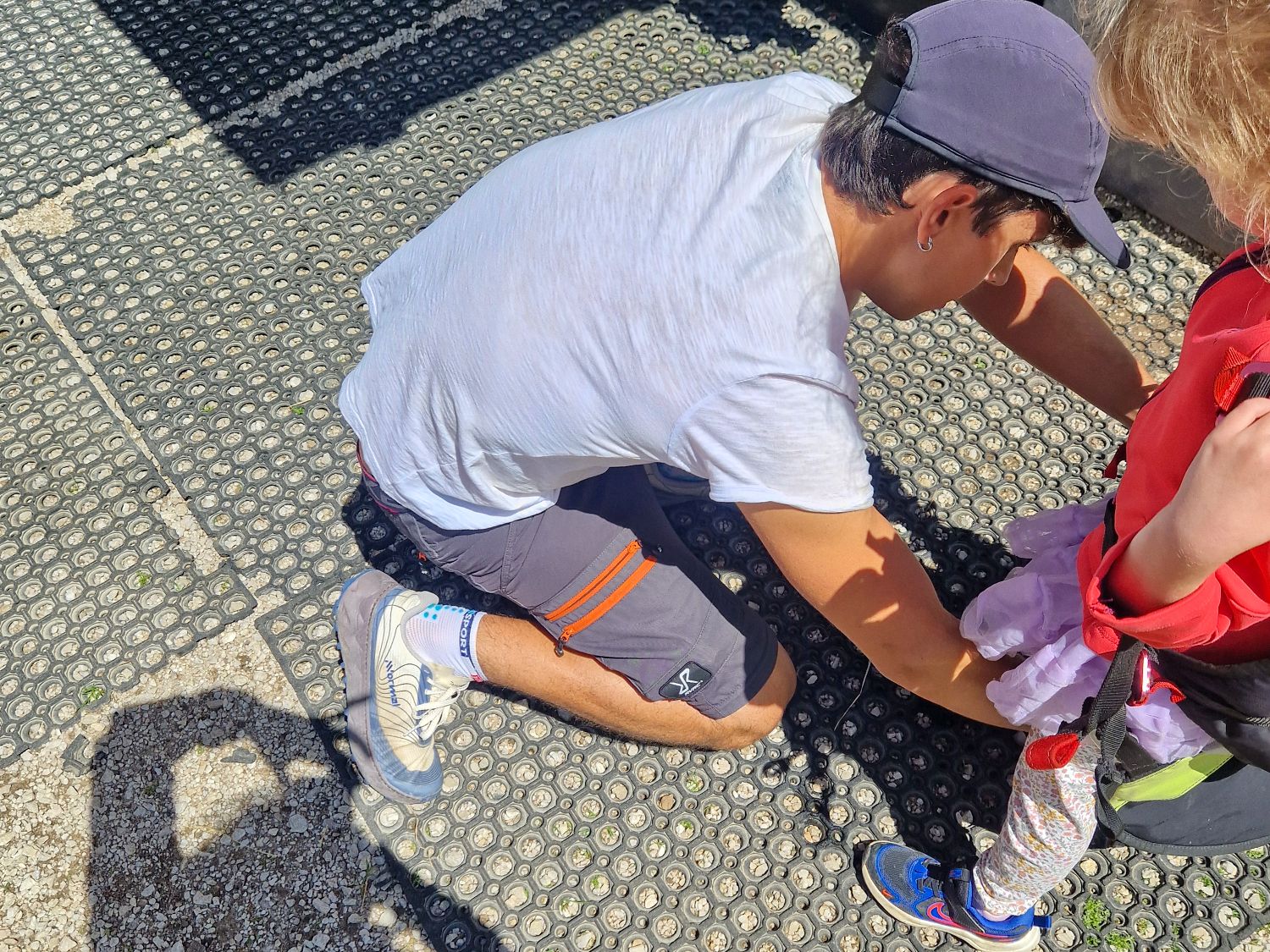 A man in a white shirt and gray shorts kneels on a rubber mat outdoors, helping a child in a red jacket and purple tutu. The sun casts strong shadows, including one of the person taking the photo.