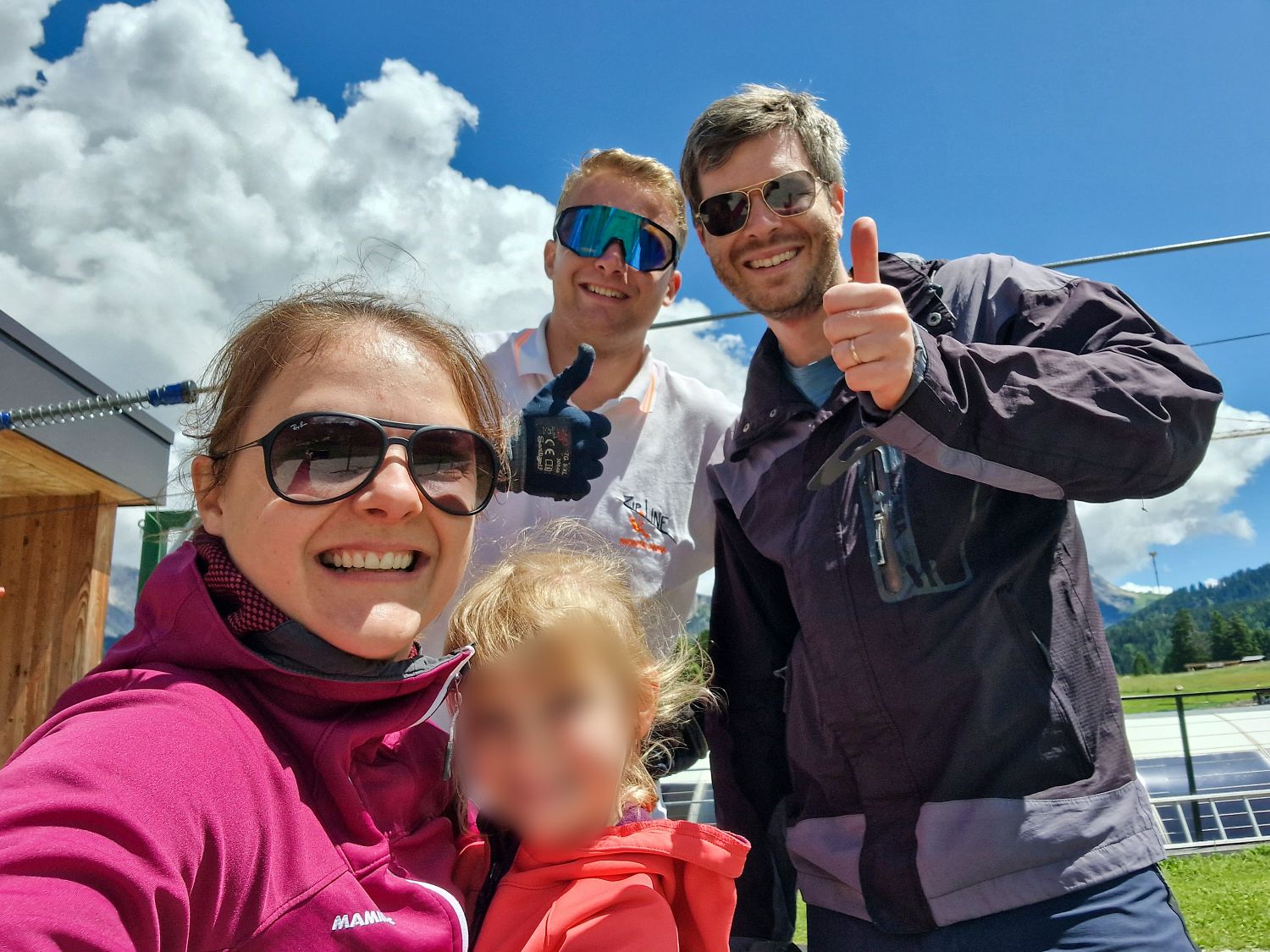Four people outdoors on a sunny day, three adults smiling and wearing sunglasses, one giving a thumbs up. The child in front is blurred. Blue sky with clouds and green hills in the background.