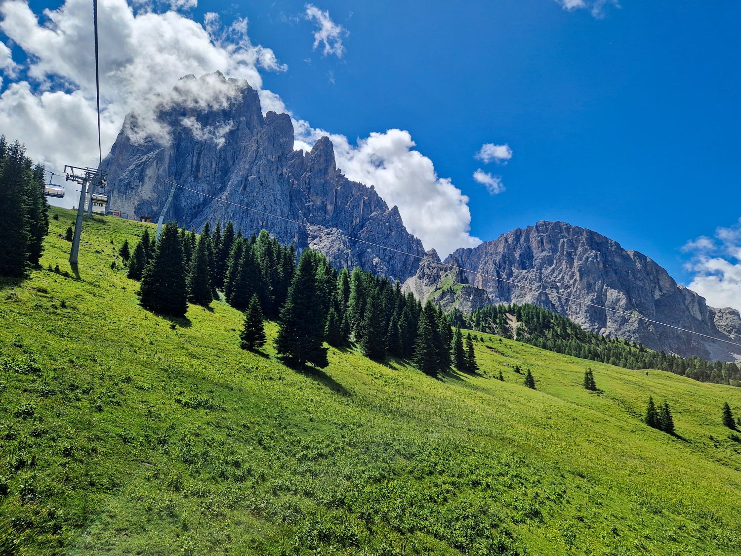 A lush green hillside with scattered pine trees under a blue sky, rugged mountain peaks in the background, and a ski lift running across the slope. Fluffy clouds partially cover the mountain tops.