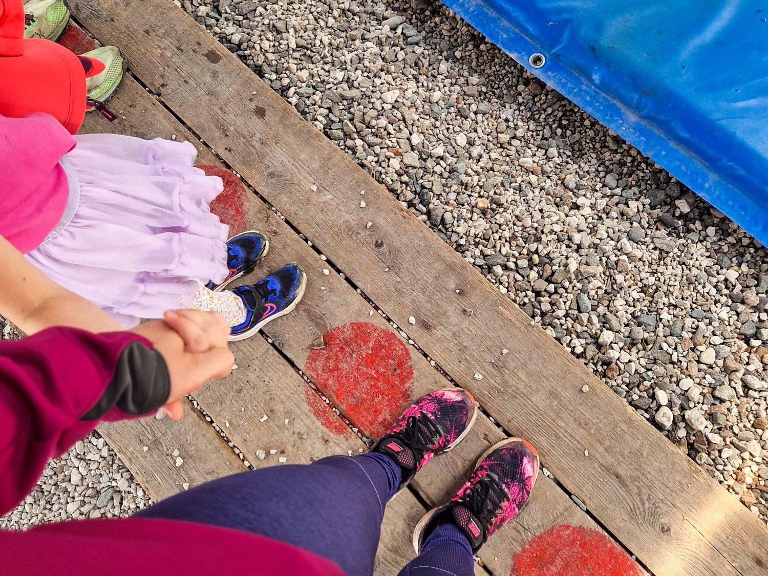 Two people holding hands stand on wooden planks marked with red circles, surrounded by gravel. One wears a pink skirt and blue shoes; the other wears purple leggings and pink-patterned sneakers. A blue tarp is visible above.