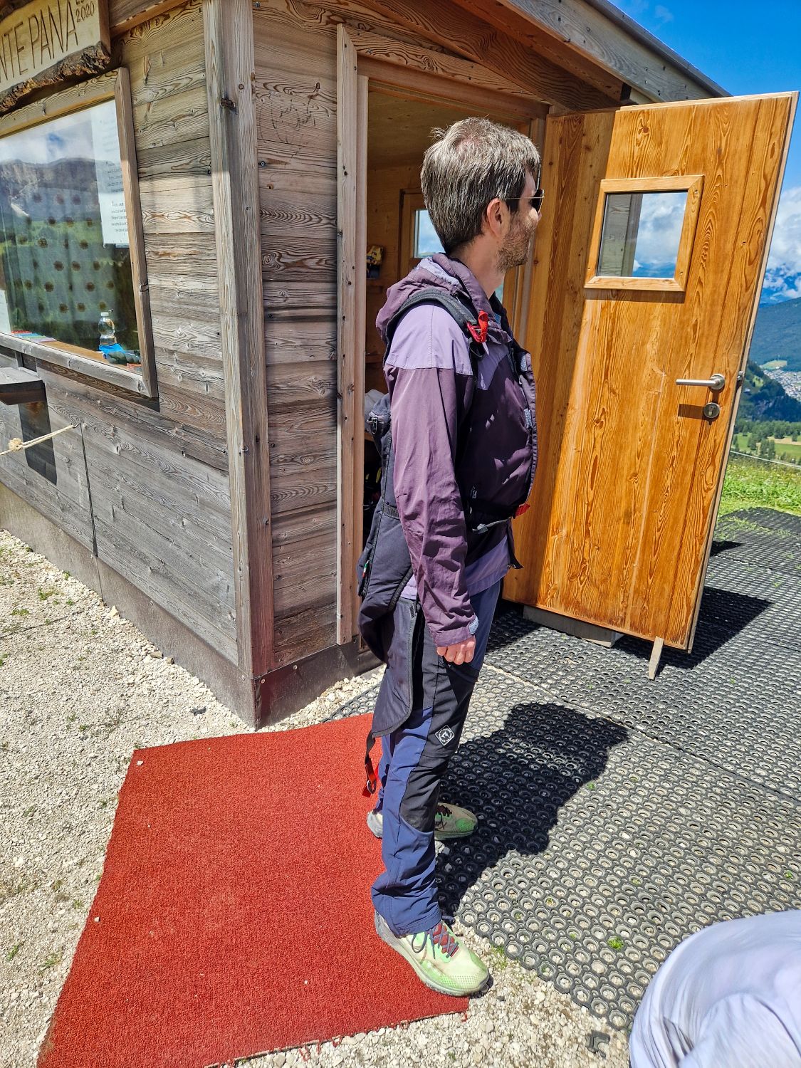 A man in hiking gear stands on a red mat outside a wooden cabin with an open door, looking away. Sunlight casts shadows, and mountains and blue sky are visible in the background.