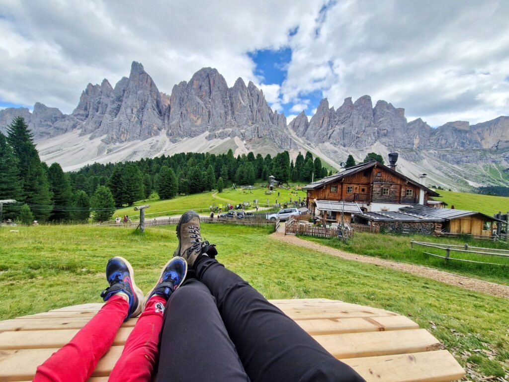 Travel Consultation 1 Two people lying on a wooden bench in a grassy field with a rustic cabin, pine trees, and dramatic mountain peaks in the background under a partly cloudy sky.