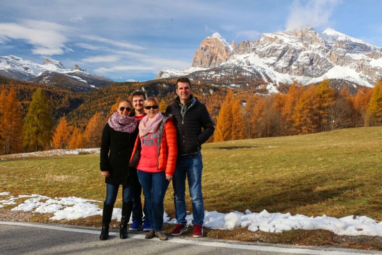 Four people stand together outdoors in front of snow-dusted mountains and autumn-colored trees, wearing jackets and scarves, enjoying a sunny day with patches of snow on the ground.