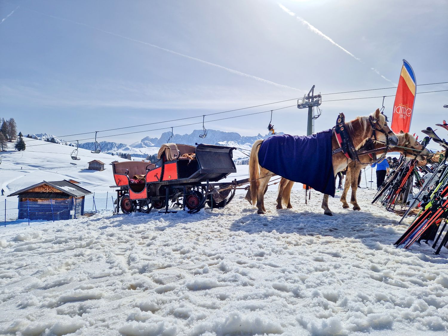 Things To Do In The Dolomites In Winter: Ski And No Ski 14 Two horses wearing harnesses and blankets stand in the snow beside a red carriage near a ski area. Skis are lined up on racks to the right while chairlifts, small wooden huts, and distant mountains sit under a bright sunny sky.